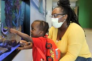 Adult and child looking at aquarium in Shawn Jenkins Children's Hospital. Child is pointing at the glass. Adult is holding child and wearing a mask. 