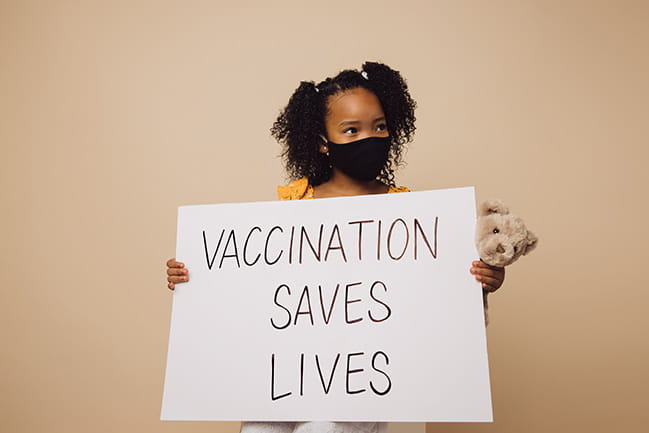 Young girl wearing a face mask holds a sign saying Vaccination Saves Lives