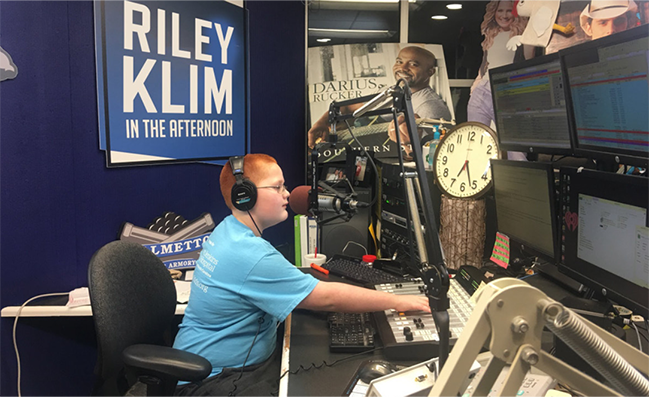 A person in a radio studio sitting at a control panel, wearing headphones, with a sign that reads "RILEY KLIM IN THE AFTERNOON" on the wall. 