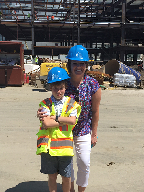 A woman and a young boy pose in hard hats at a construction site. The woman is smiling. The boy has crossed his arms and looks as though his mother forced him to pose. 