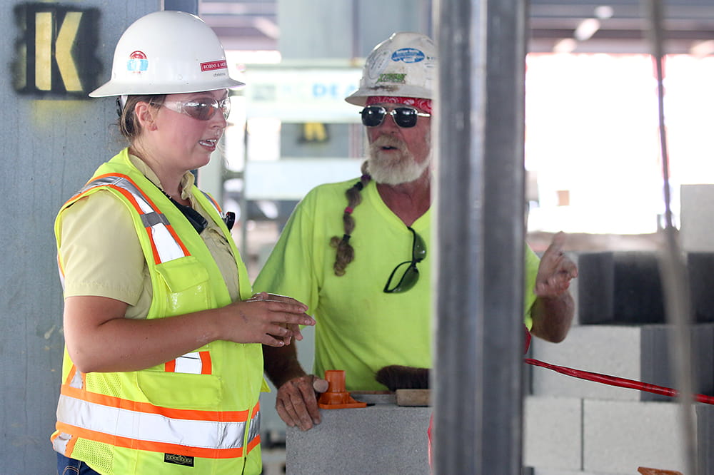 A young woman in a safety vest and hard hat talks to an older male in a hard hat inside the construction site