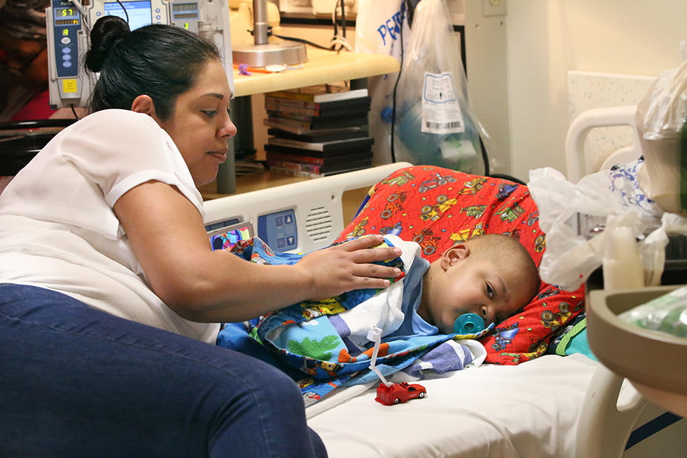 A mother sits on a hospital bed and leans over her young son, who lays on his side. A tiny fire truck is on the bed