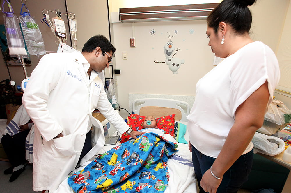 A doctor leans down to touch a small boy in a hospital bed as his mother looks on
