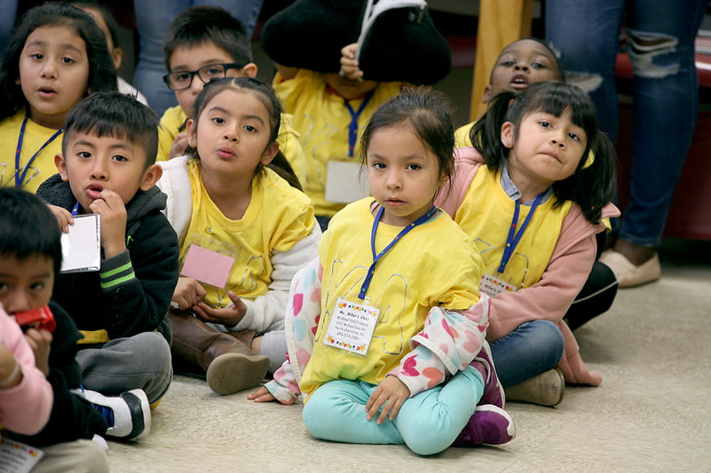 A group of kindergartners sit on the floor to listen to a presentation