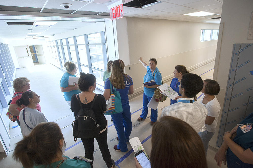 A group stands in the hallway of the not-yet-finished hospital. Brown paper is taped to the floor. Many of the people hold maps. 