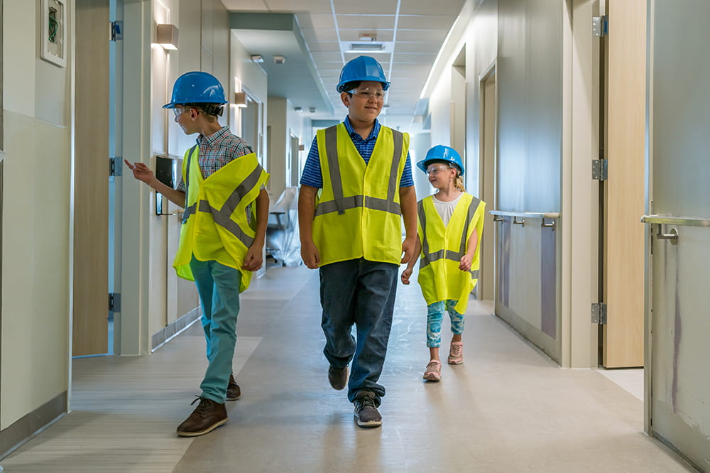three children in hard hats and adult-sized yellow vests walk through the halls of the nearly-complete hospital