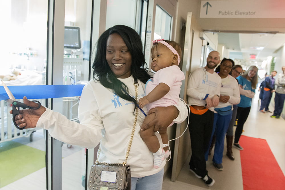 woman cuts a blue ribbon outside a patient room while holding baby