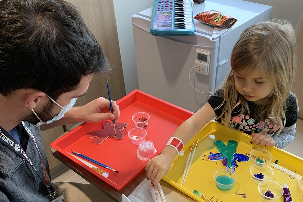 A little girl paints flat Christmas tree ornaments with a medical student
