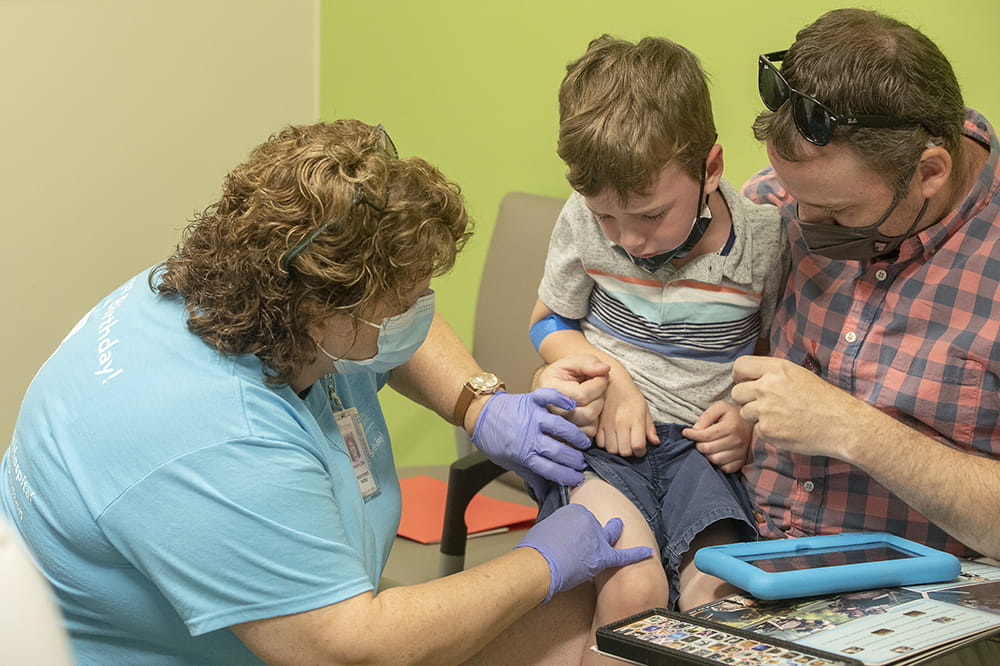 Nurse Karen Hawkins puts a Band-Aid on Ethan Appleby after his first dose for the Moderna vaccine in the dosing clinical trial for children from 2 to 5 years old while his father, Scott Appleby, holds him.