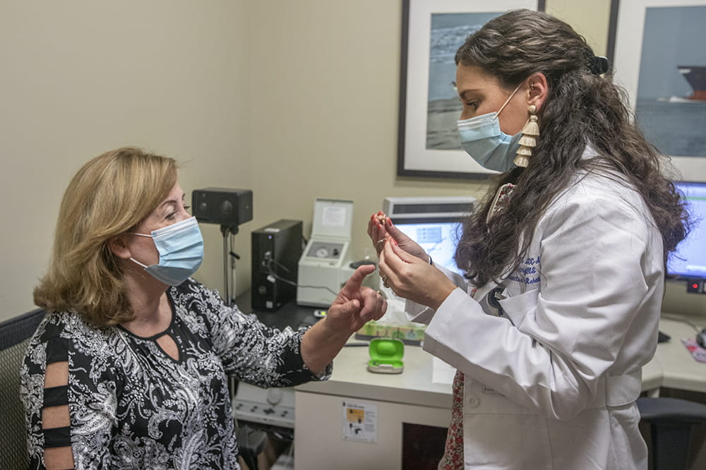 a woman sitting points to a device in the hand of a standing female audiologist