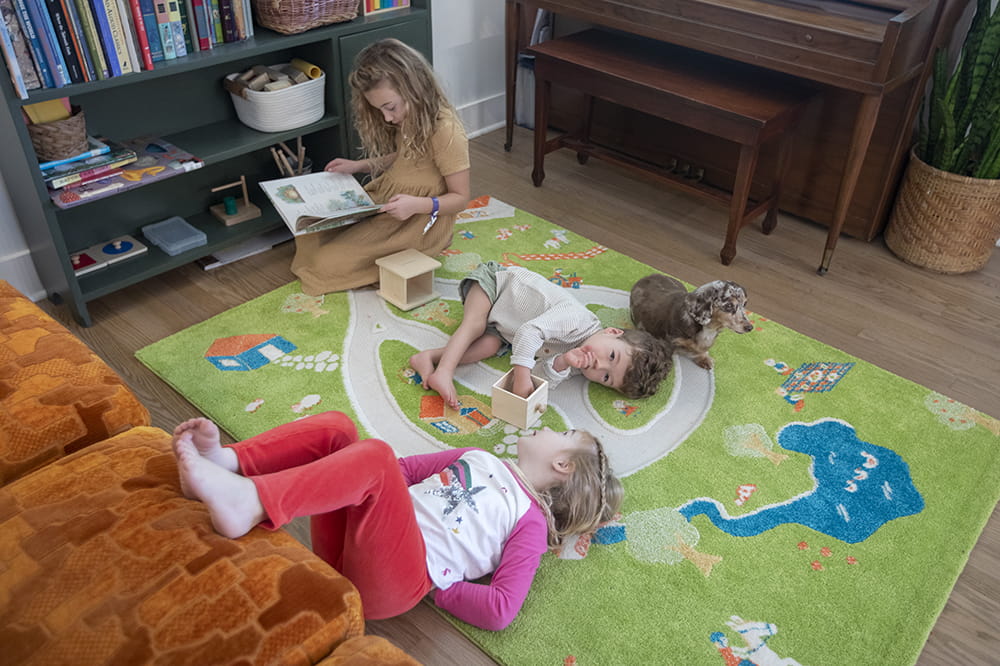 three children lounge on a colorful rug of the countryside surrounded by a piano and bookshelf. the oldest girl looks at a book while the younger girl and the boy lay on the floor being silly
