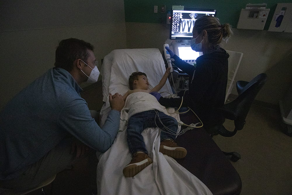 in a darkened exam room, a little boy lays on a table with his father holding his hand on one side. on the other side, the tech lets him squeeze gel onto the wand while the computer monitor glows in the darkness