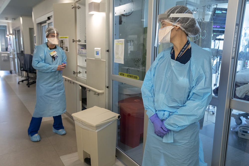 two women in blue scrubs, gloves, gowns, hair nets, masks and face shields stand outside a patient room 