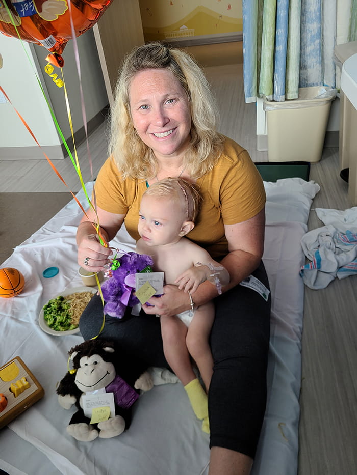 a woman sits on a mat on the floor of a hospital room, holding balloons in one hand and a baby in a diaper between her legs. He has a large scar on his head
