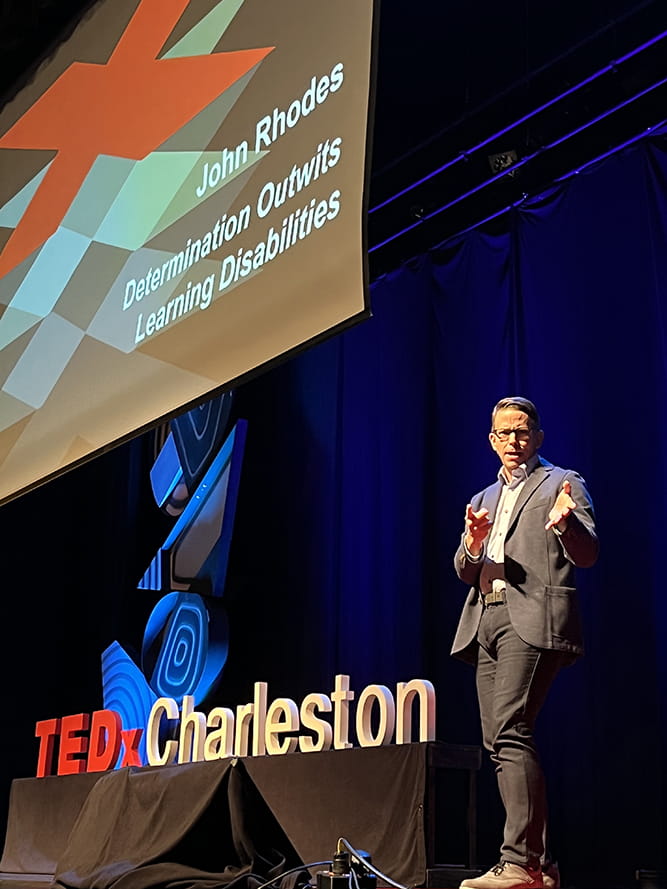 a man stands on a stage beneath a large screen with a red X and in front of a sign that says TEDxCharleston