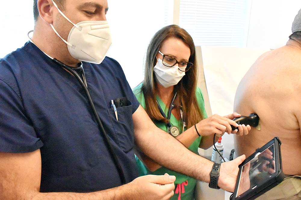 Dr. Rami Zebian holds a smart device while a woman next to him uses a Butterfly ultrasound device on a man's bare back.