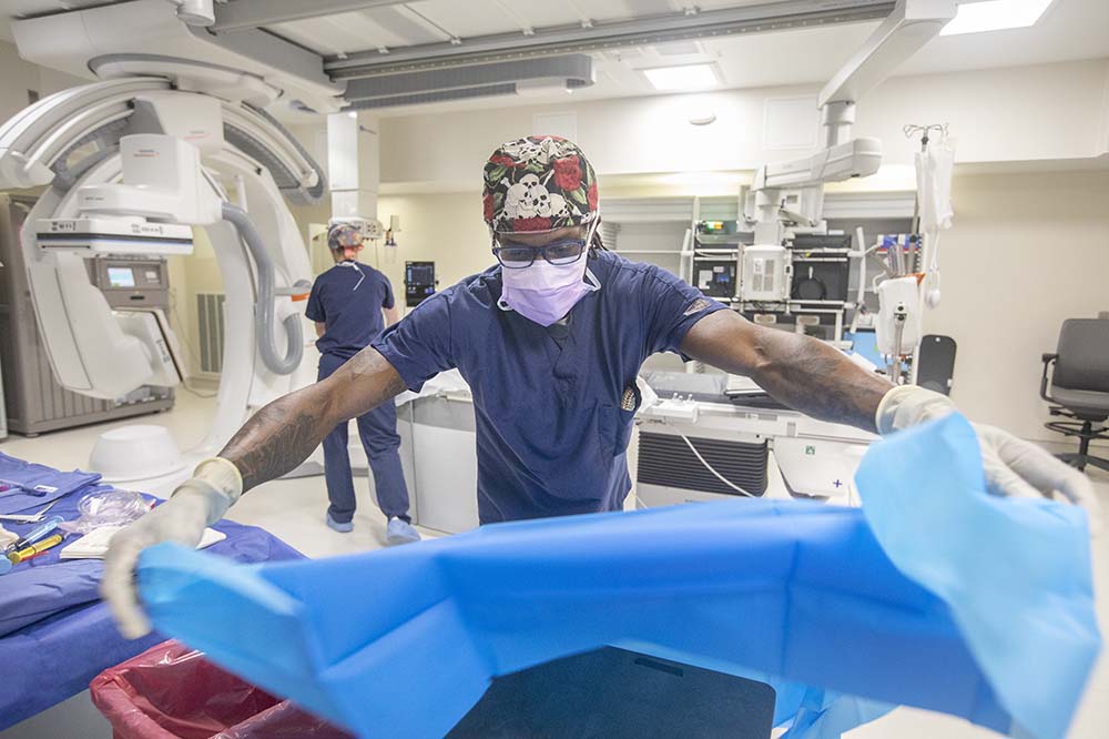 Tech wearing a mask with his hair covered stretches his arms as he spreads a blue tarp in a new endovascular suite.