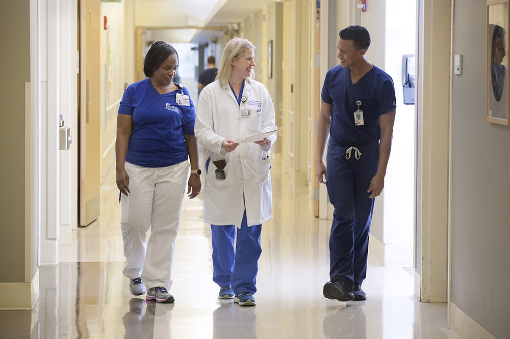 Left to right: Pamela Bowers, BSN, RN  Dr. Heather Simpson M.D.  Keeland Williams, COM student. Three people smile while walking in a hall.