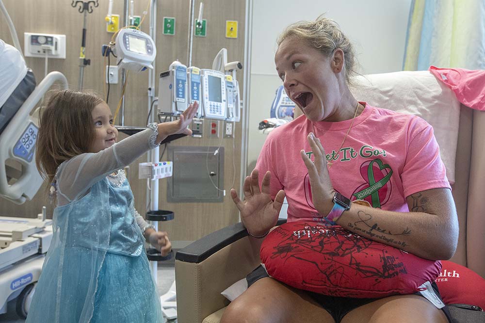 Natalyn Mann holds up a hand to her aunt Sara Cathey to freeze her. Natalynn is wearing a blue princess dress. Sara is wearing a bright pink t-shirt. Sara looks delighted.