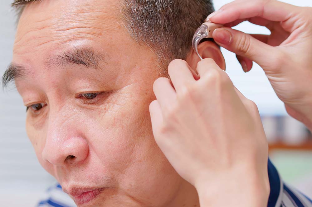 Man looks down while woman's hands put hearing aid on his left ear.