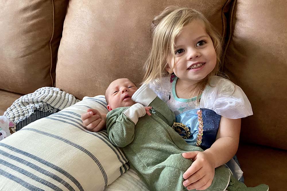 Liam and Aurora Robertson. Liam, a baby, is lying in Aurora's lap. He's wearing a green onesie. Aurora is wearing a princess dress and smiling.