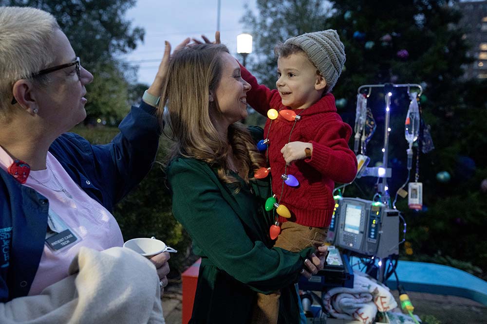 Smiling mother holds her son Luke at the Angel Tree lighting ceremony. A cart with IV bags stands beside then.