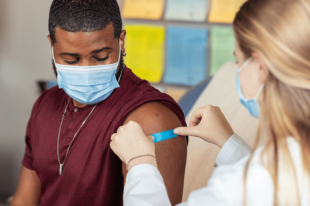 Man wearing a surgical mask looks at woman putting bandaid on his arm where he just got a flu shot.