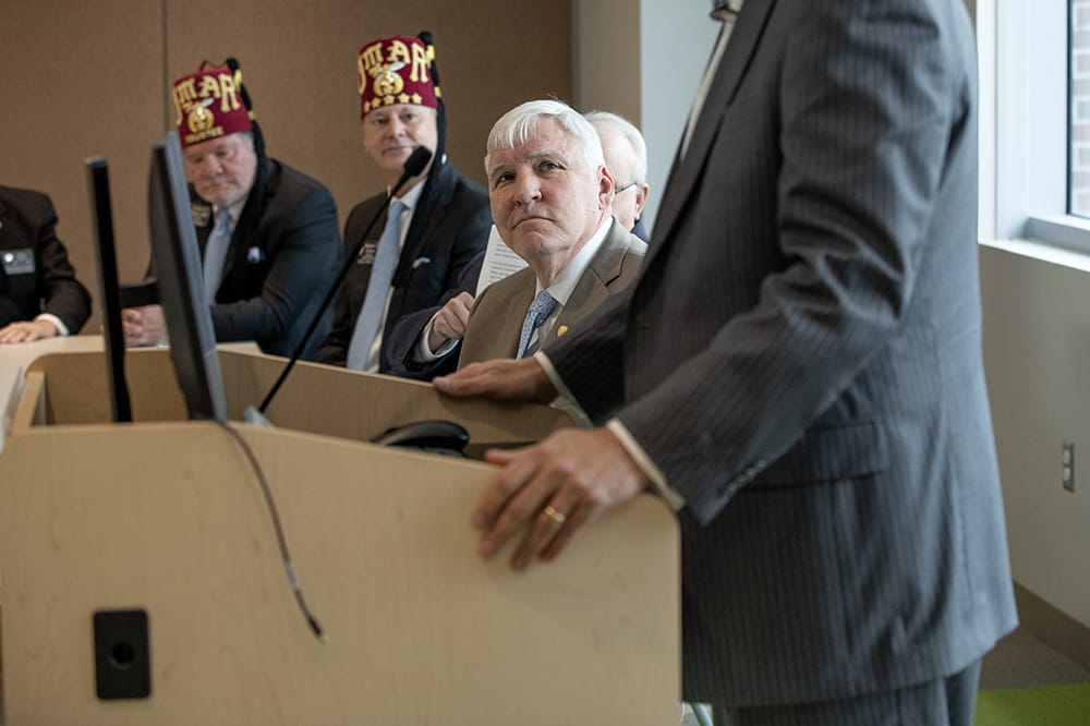 MUSC President Dr. David Cole is seated as he listens to a Shriner at a podium speak. They are in a conference room.