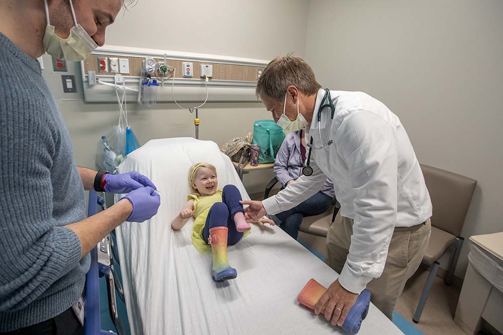 Little girl lying on bed laughs while doctor looks at her feet.