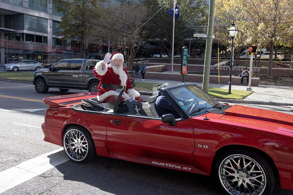 Santa waves while sitting on the back of a red convertible.