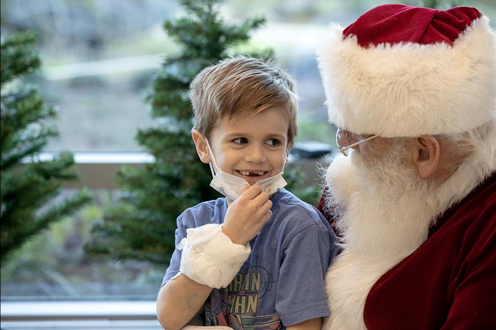Little boy with missing front teeth smiles as he pulls down a face mask to talk to Santa.