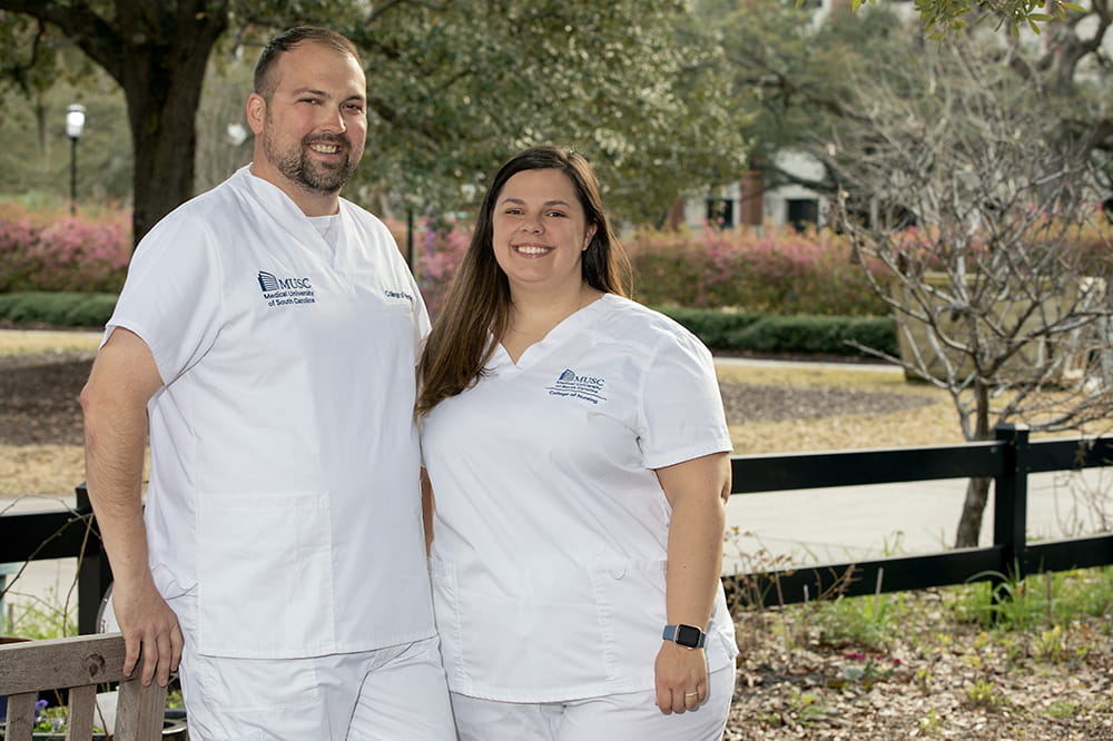 A man and a woman in white scrubs stand outside in front of a wooden fence
