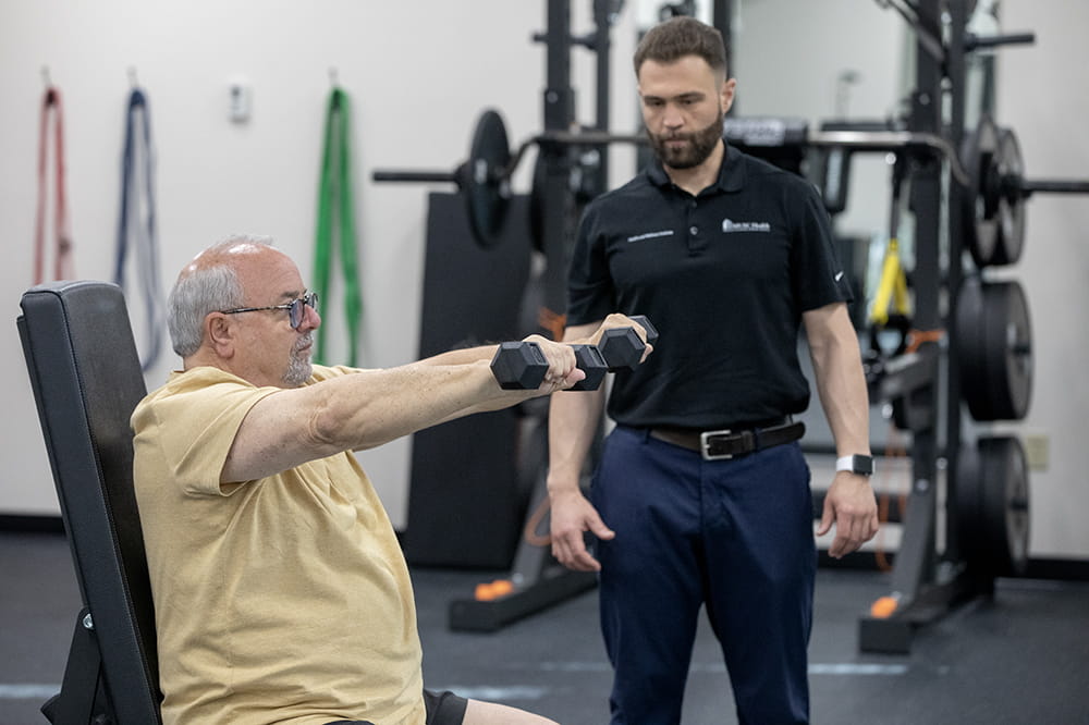 Man seated lifts hand weights while his coach watches. They are in a workout room.