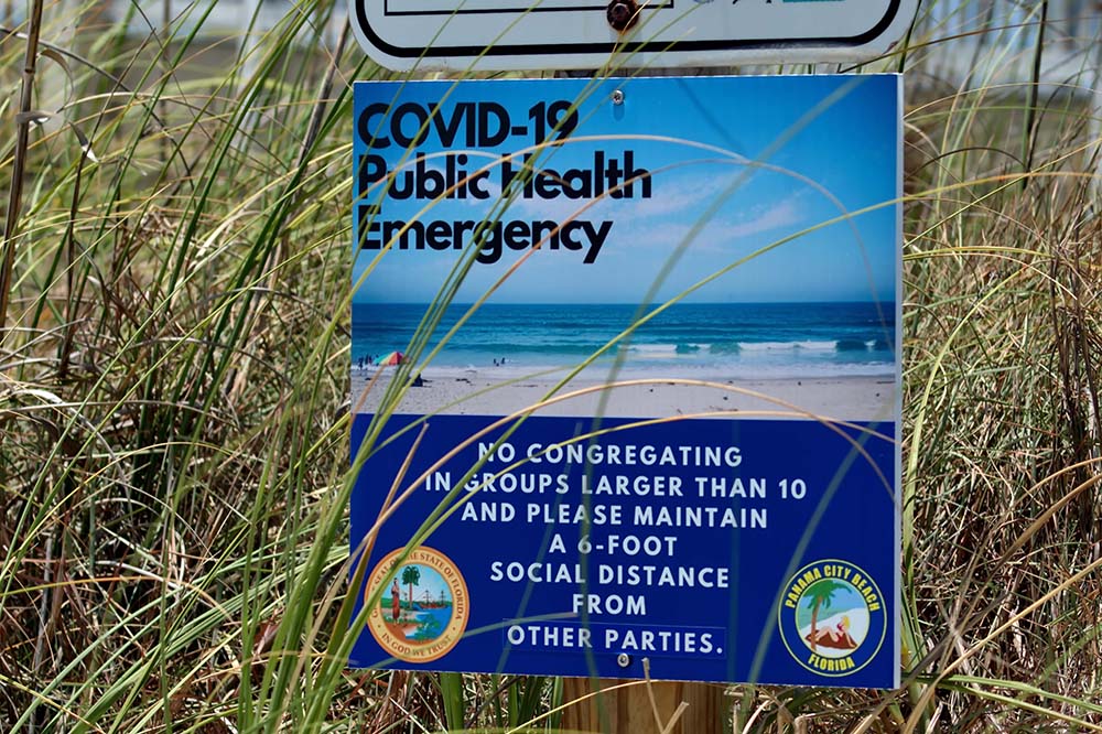 Blue sign against backdrop of sea grass. The sign says COVID-19 Public Health Emergency. No congregating in groups larger than 10 and please maintain a 6 foot social distance from other parties.