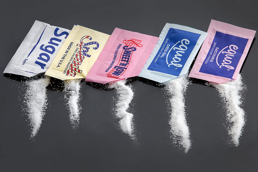 Packets of sugar, Splenda, Sweet'N Low, Equal and Equal are linked up with sweeteners pouring out of them.