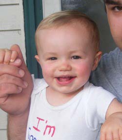 Little boy wearing a onesie is smiling while being held by an adult. They are holding hands.