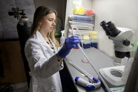 A young woman in a white lab coat and blue gloves holds a tube in a lab