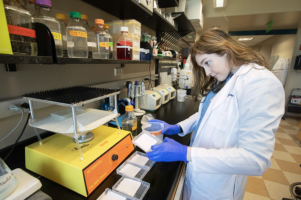 A young woman in a white lab coat looks at a device inside a lab