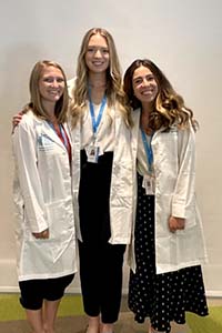 Three young women with long hair wearing white doctors' coats smile for a photo.