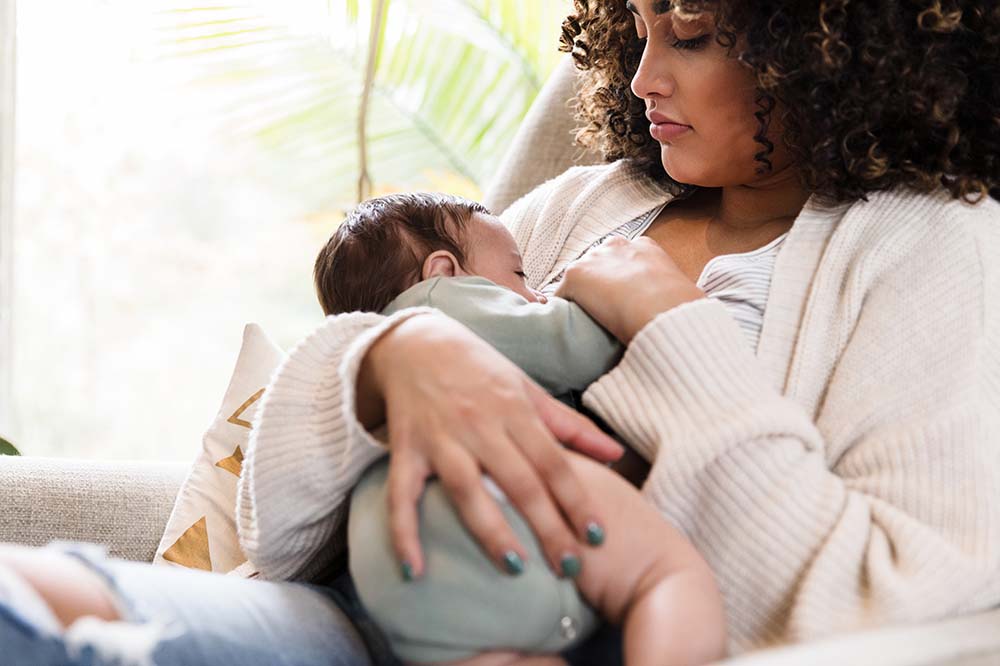 Woman with dark, curly hair breastfeeds a baby. The woman is wearing a beige colored sweater. The baby is in a gray onesie.