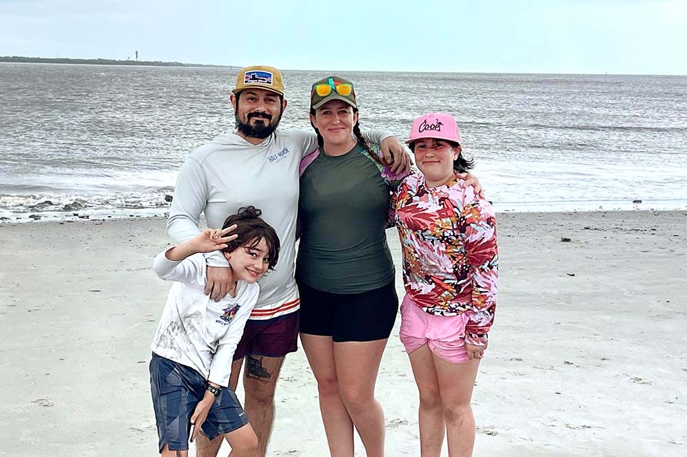 Family of four smiles at the beach. The parents and daughter are wearing caps. The son is leaning on the father.