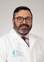 Man with a beard and glasses smiles for a portrait. He is wearing a white doctor's coat with the logo for the children's hospital on it.