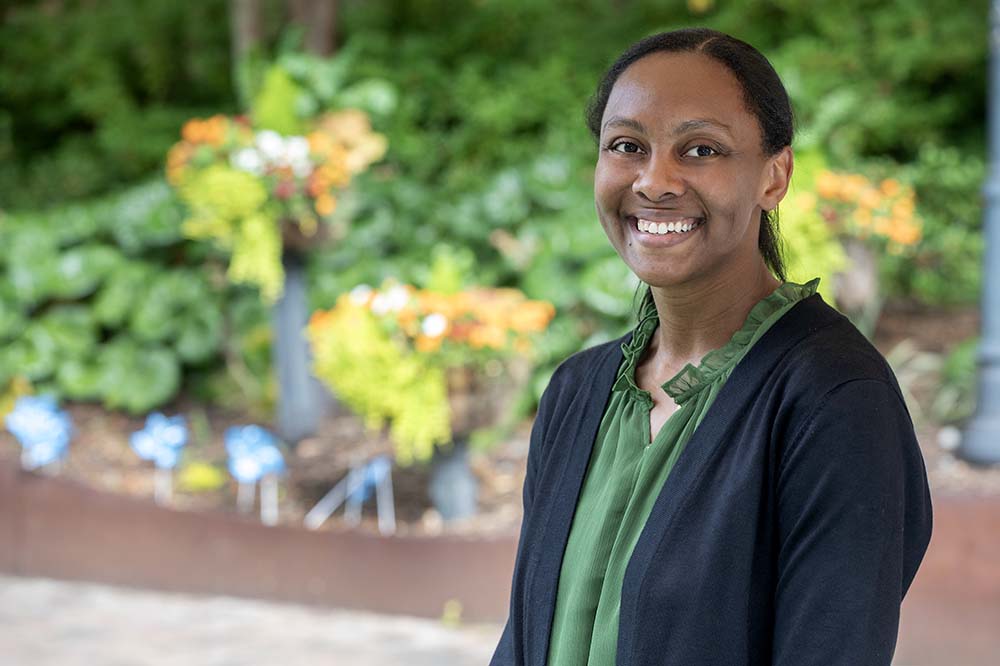 Woman with her hair pulled back smiles in front of flowers. She is wearing a blue blazer and green blouse.