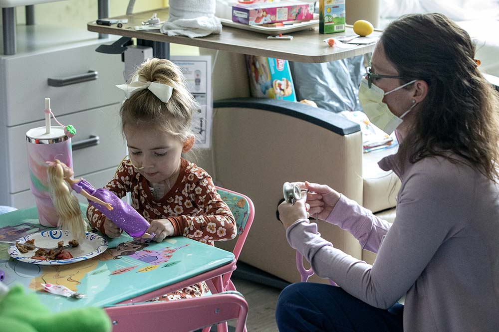 Girl looks at purple toy as a woman beside her wearing a surgical mask talks to a third person off camera.