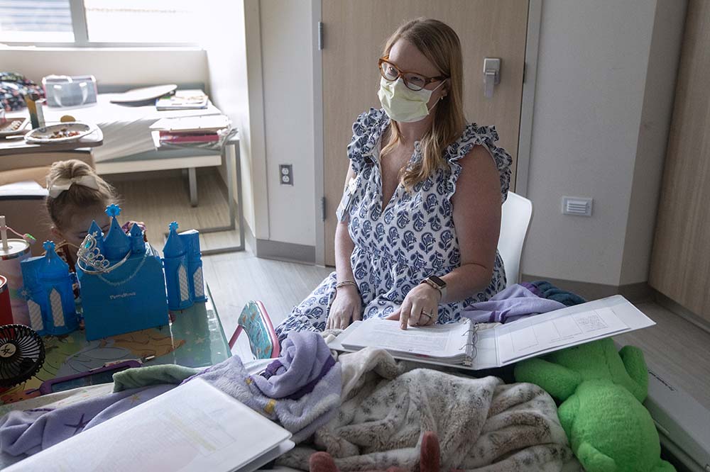 Woman in a sleeveless white dress wearing a surgical mask talks with a notebook open in front of her. A girl sits at a table to her side.