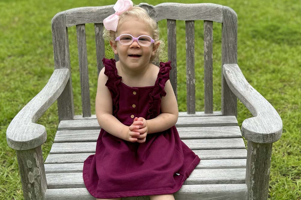 Girl with blonde curly hair sits on a bench. She is wearing a maroon velvet dress, pink glasses and a bow in her hair.