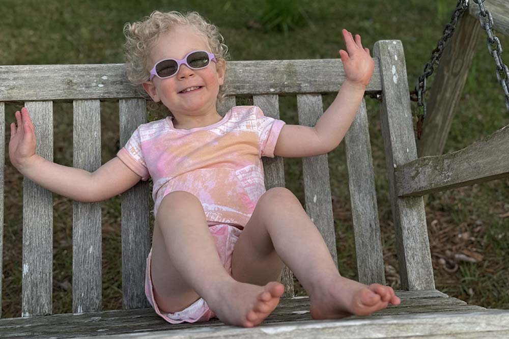 Girl in pink shorts and top smiles while sitting on a wooden bench.