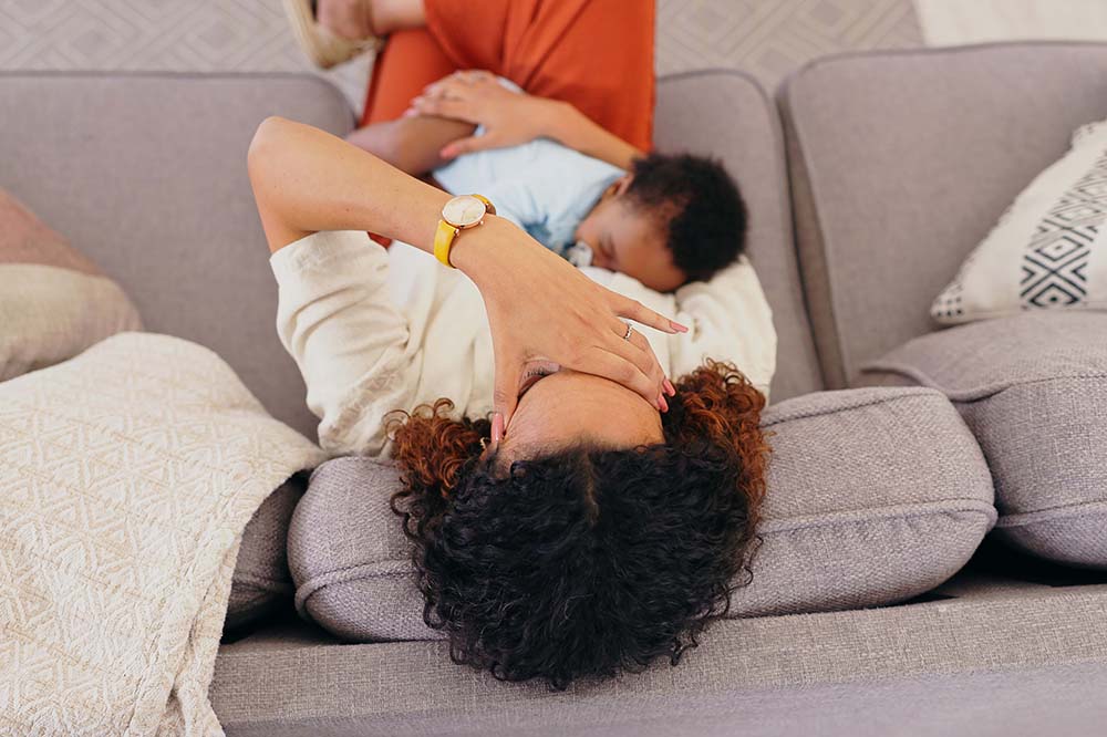 Woman with dark curly sofa lies upside down on a sofa with a baby on her chest. Her hand is covering her face.