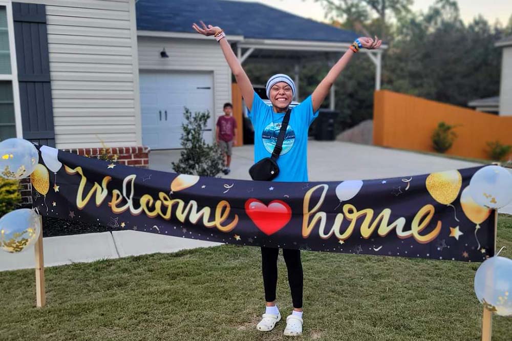 A girl wearing a cap throws her arms up behind a sign that says welcome home.