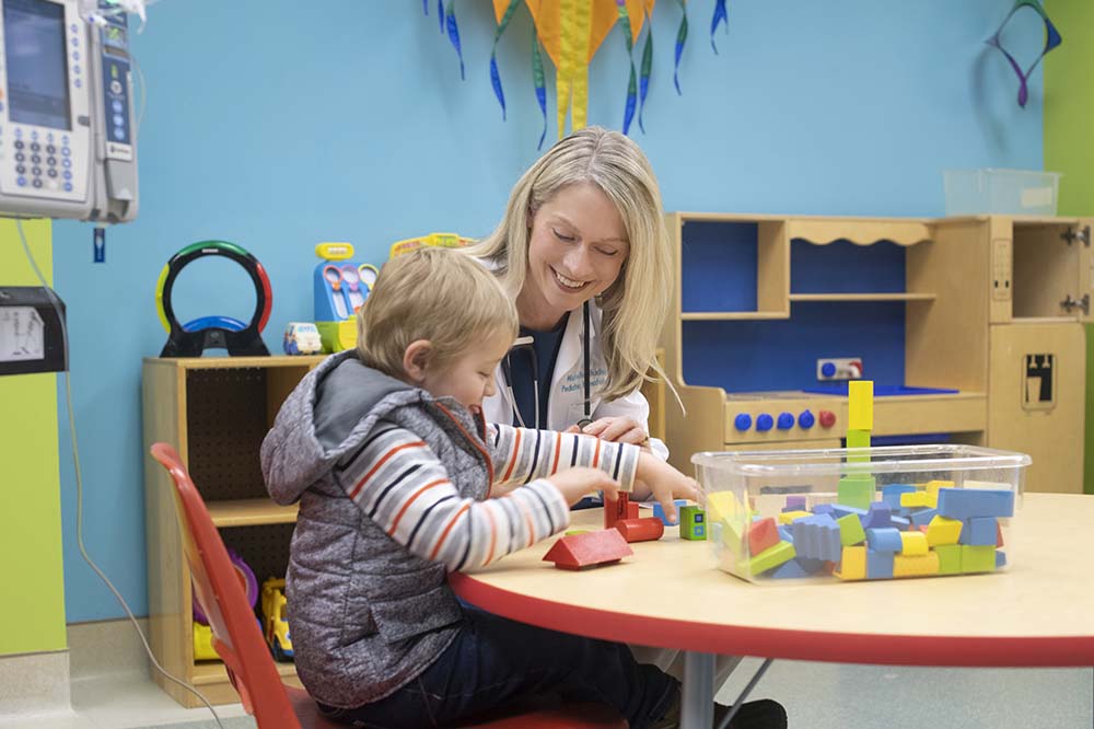 A doctor with long blonde hair sits with a child at a table with toys on it.